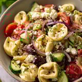 Schüssel mit bunter Nudelsalat, Tomaten, Gurken, Zwiebeln und Feta.