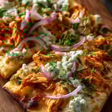 Eine halbe Pizza mit Chicken, Onion, Pink Pepper und Blue Cheese auf einem Holzblock.